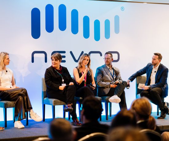 The photo of a panel discussion at a Nevro conference, featuring five professionals seated on a stage. One woman in the center is speaking into a microphone, while the others listen attentively. The backdrop displays the Nevro logo, and the audience is visible in the foreground.